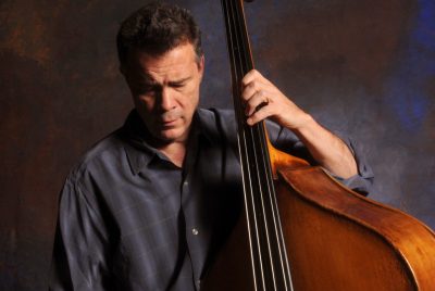 Musician Edgar Meyer holds onto a large stand-up bass, with his fingers placed on the frets he looks down to the floor.
