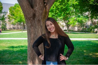 A portrait of Betsabeth standing in front of a tree trunk.