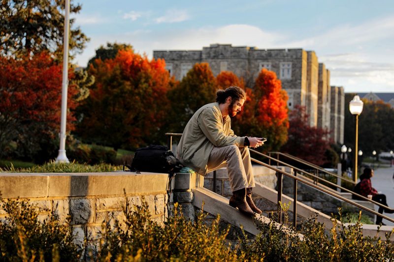 Male student sitting outside on a stone wall with autumn foliage